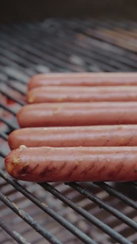 A vertical closeup of Several hot dogs with light grill marks are lined up on a dark grill grate.