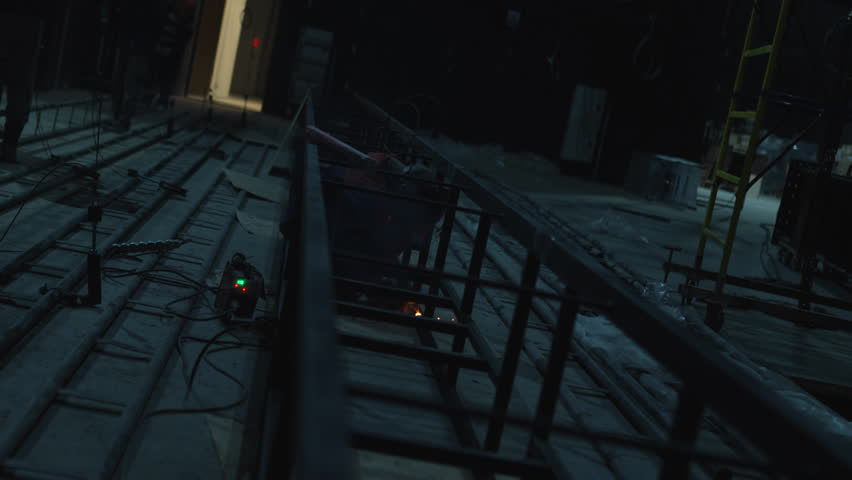 Welder carefully works on metallic construction in dim hall. Dark workshop filled with metallic framework glows with welding sparks