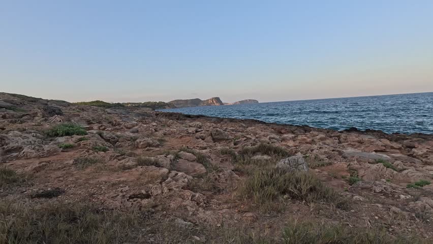 Ibiza rocky coastline with dry grass and Mediterranean waves under clear evening sky, natural seascape capturing island beauty, peaceful summer landscape and travel destination atmosphere.