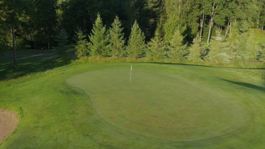 Close-up aerial view of the putting green at Avoti Golf Course, surrounded by trees and neatly trimmed fairways in the early morning sunlight.
