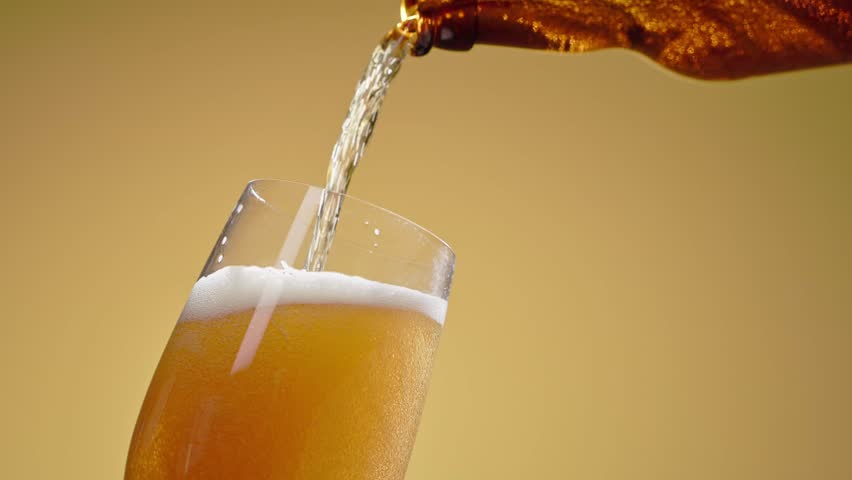 Cold craft light beer pouring in glass from bottle, slow motion close up. Stream wheat malt beverage flowing in clear goblet making dense bubbling foam. Froth heady drink foaming on yellow background