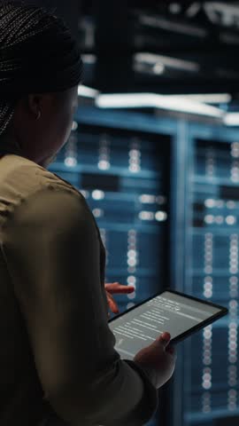Vertical video Close up of data center programmer doing data analysis on tablet to upgrade infrastructure. African american woman in server room using device to manage storage systems datasets