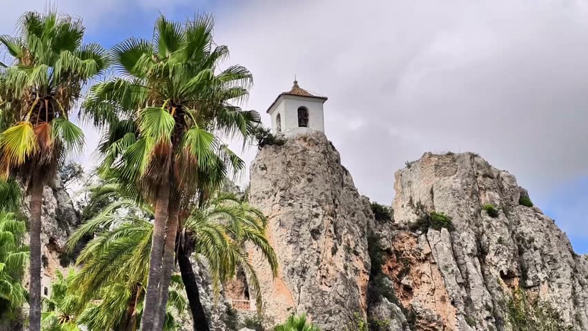A beautifully designed chapel perched high atop a rocky cliff, elegantly framed by lush palm trees beneath a cloudy sky