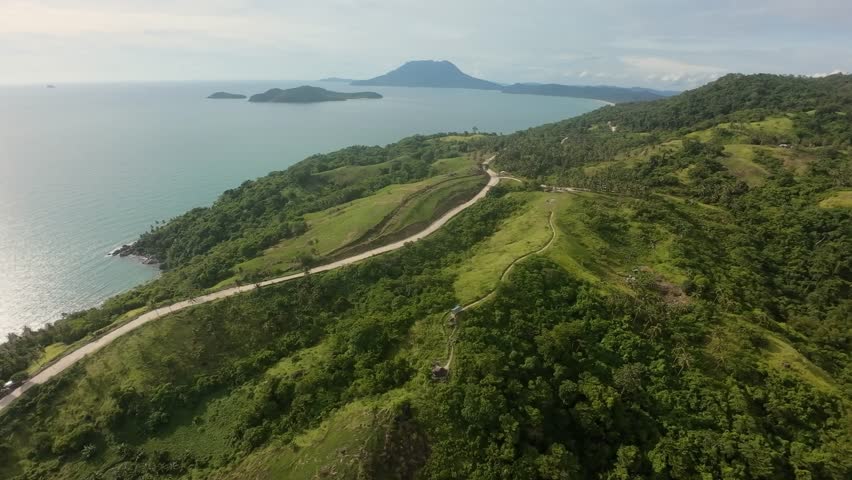 Minute-long FPV run with banked turns above Bato ni Ningning: curving ridge road, palm-studded hills and the arc of Long Beach in San Vicente, with afternoon sun glint on turquoise sea.
