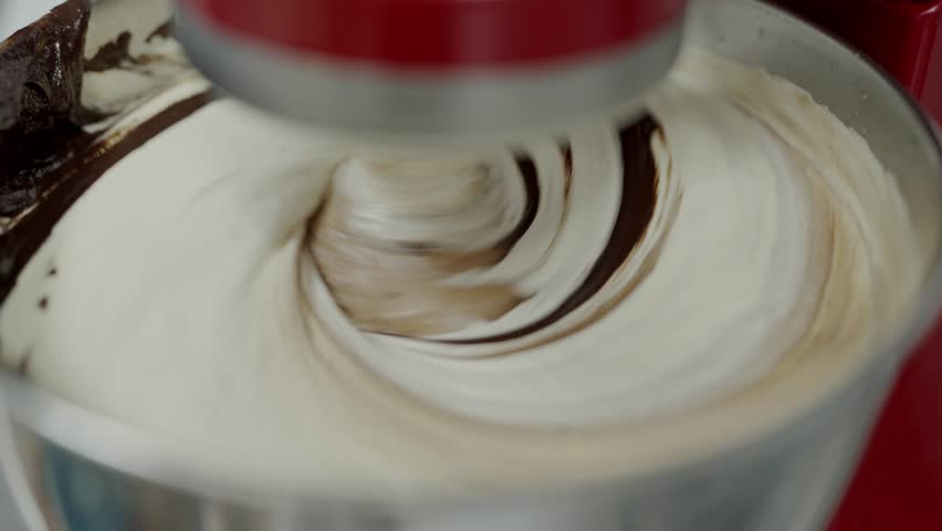A detailed view of cream being whipped in a stainless steel bowl by a red stand mixer, highlighting the texture and motion. Close-Up of Cream Whipping Process in Red Mixer, Kitchen Setting
