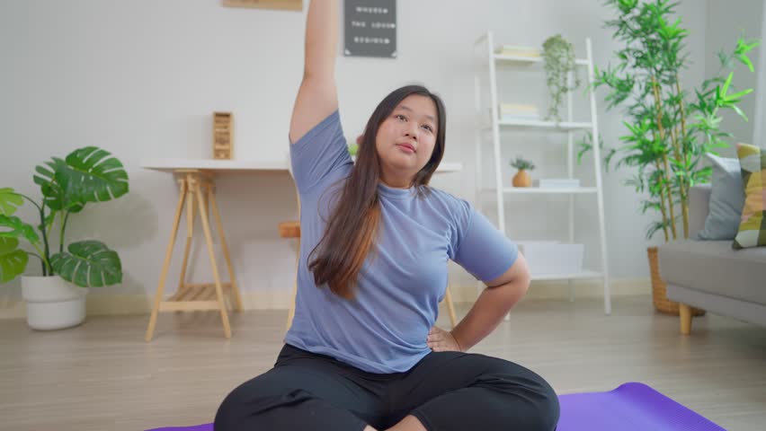 Asian plus-size woman exercising to lose weight in living room at home. Attractive young curvy female spend weekend leisure time doing yoga workout and stretching body to improve health and fitness.