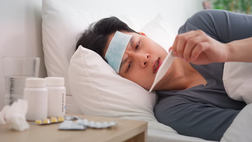 Asian young man lying down on bed in bedroom, looking sick and unwell. Attractive man having fever gel pads on his forehead, suffering from fever and headache after waking up in the morning at home.