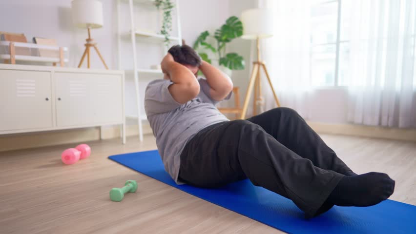 Exhausted Asian plus-size man workout and try to sit up on yoga mat. Attractive young curvy male spend leisure time exercise and doing cardio to improve health and lose weights in living room at home.