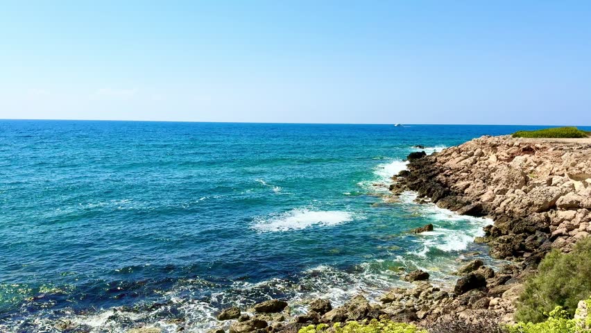 Public beach, summer, beautiful sunny weather. Picturesque beach, waves crashing on the sandy shore. Sea waves beat against the rocks. Cyprus Island, umbrellas, tourist boats, Mediterranean Sea.