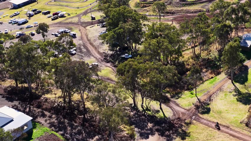 Drone captures dirt bike rider on winding forest trail in daylight, parked vehicles visible nearby.