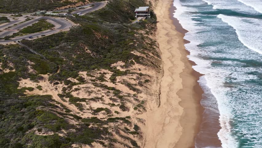 Drone glides above Portsea coastline, capturing waves, sandy beach, and modern lifeguard station