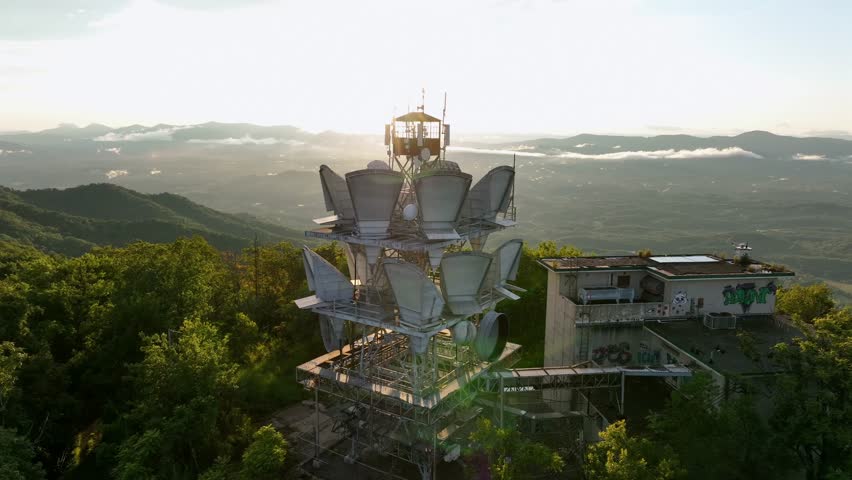 Aerial view of American radio tower on top of green mountains during sunrise. Graffiti on wall of building on peak. Beautiful landscape and clouds in valley.