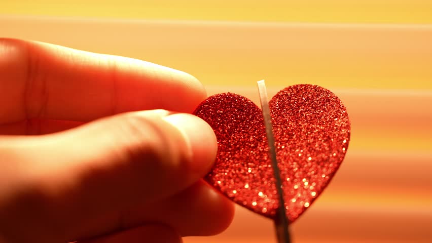 A heartbroken person uses scissors to cut a red paper heart in backlight, sunlight streaming through blinds behind, symbolizing emotional pain, separation, vulnerability, healing, and the letting go.
