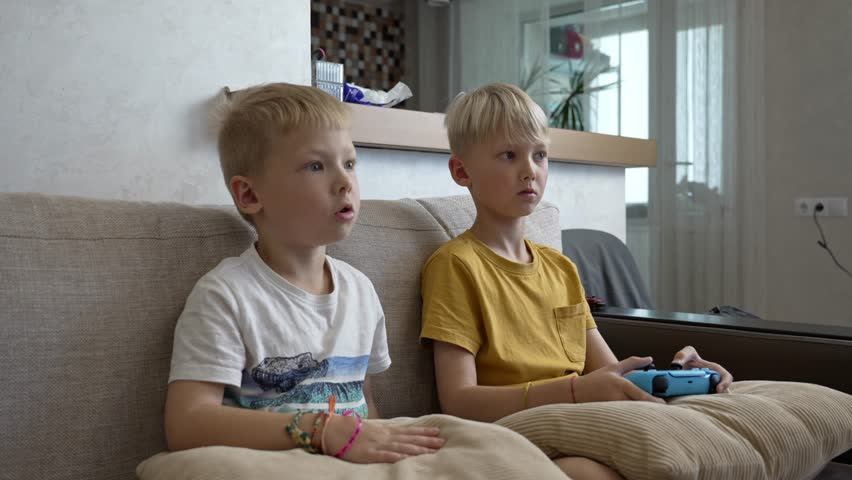 Two boys sit intently on a sofa, fully immersed in a gaming session. The sunlit room features a modern aesthetic, adding to the atmosphere of friendly competition and teamwork.