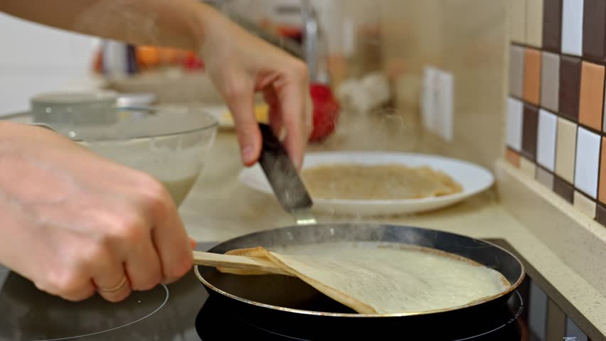 Woman chef is skillfully flipping a thin pancake in a non-stick frying pan, using a wooden spatula, creating a delicious breakfast in a modern kitchen