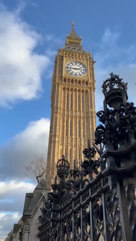 Low-angle of London's Big Ben clock tower, known as the Elizabeth Tower. Grandeur of ornate Gothic architecture and famous clock face, with foreground element of decorative wrought-iron fence.