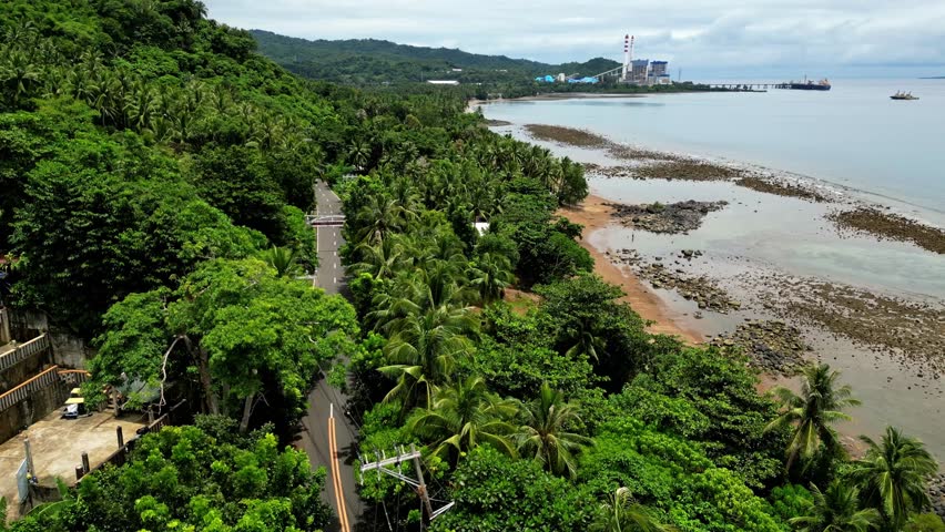 An elevated shot glides along a tropical coastline, passing lush forest, a winding road, rocky sand beaches, and calm waters beside Mauban, Quezon Province Philippines