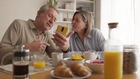 Happy senior couple enjoying breakfast and social media. Elderly couple laughing while using smartphone during breakfast at home - Powered by Shutterstock - Get 15% off with code: PIKWIZARD15