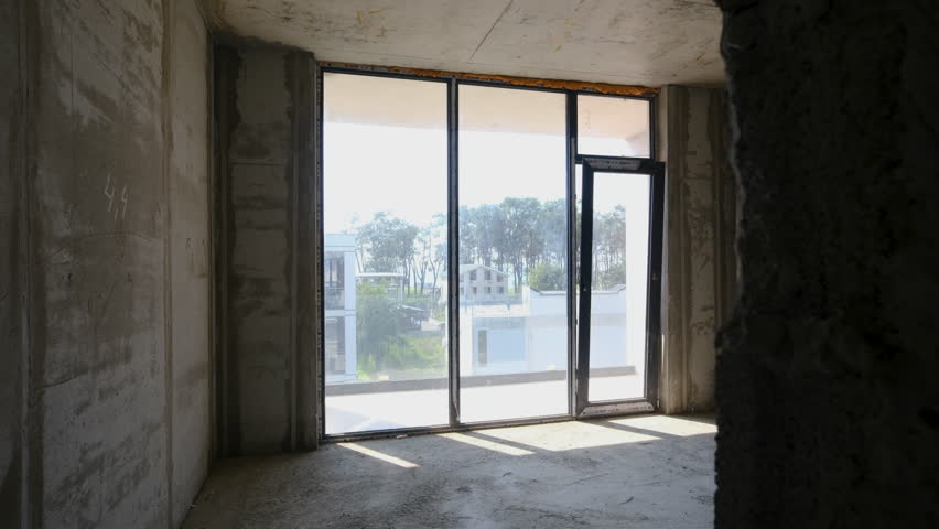 Slider shot of Interior view of a spacious, unfinished room featuring large glass windows that let in natural light, providing a view of greenery, giving an impression of future potential and openness