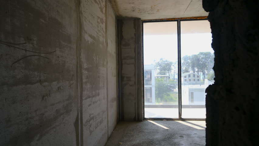 Slider shot of Interior view of a spacious, unfinished room featuring large glass windows that let in natural light, providing a view of greenery, giving an impression of future potential and openness