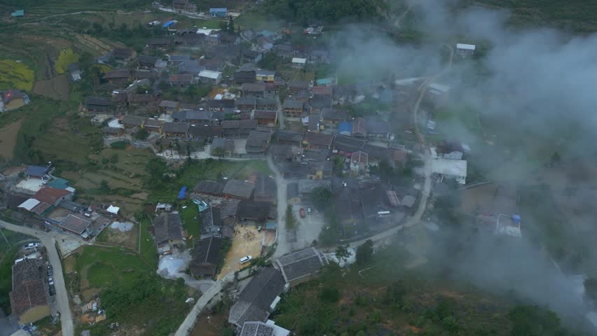 Aerial drone view of a traditional mountain village with tiled-roof houses surrounded by terraced fields and misty clouds, creating a peaceful and poetic landscape