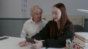 Serious senior woman in white vest sitting with teen girl in black hoodie at desk with calculator and books, symbolizing home study and support - Powered by Shutterstock - Get 15% off with code: PIKWIZARD15