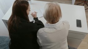 Teen girl in black hoodie writing in notebook with senior woman in white vest sitting beside at desk with calculator, symbolizing family education and home study. Overhead shot - Powered by Shutterstock - Get 15% off with code: PIKWIZARD15