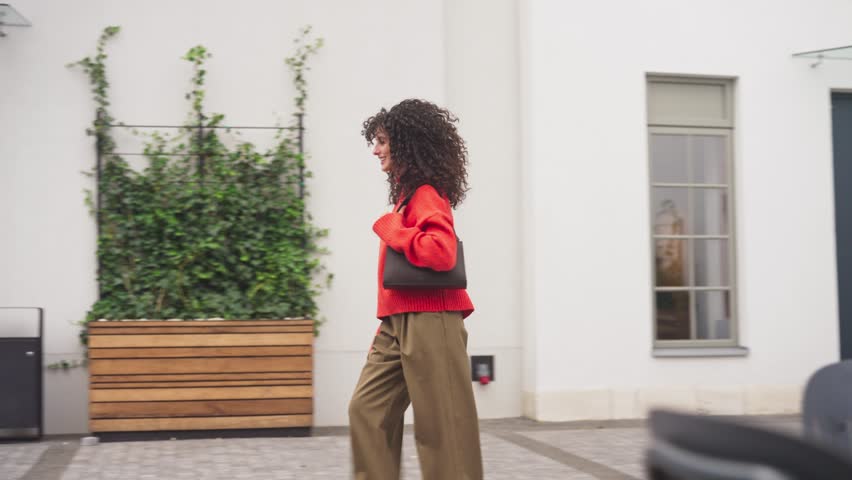 Beautiful Caucasian woman with curly hair walking outdoors near building, smiling and holding handbag. Female moving with confident step, enjoying city street lifestyle outside.