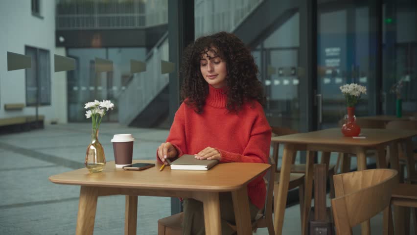 Curly haired Caucasian woman in red sweater reading book at wooden table with coffee cup, smartphone and flowers in vase near window. Female turning page while studying or relaxing indoors.