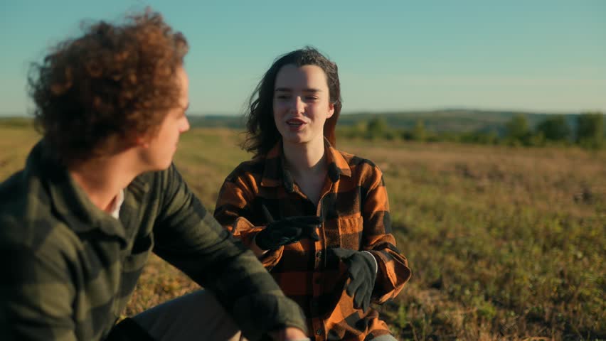 A young man and woman dressed in plaid flannel shirts squat in a farm field, talking and smiling while enjoying teamwork and connection in a peaceful rural landscape.