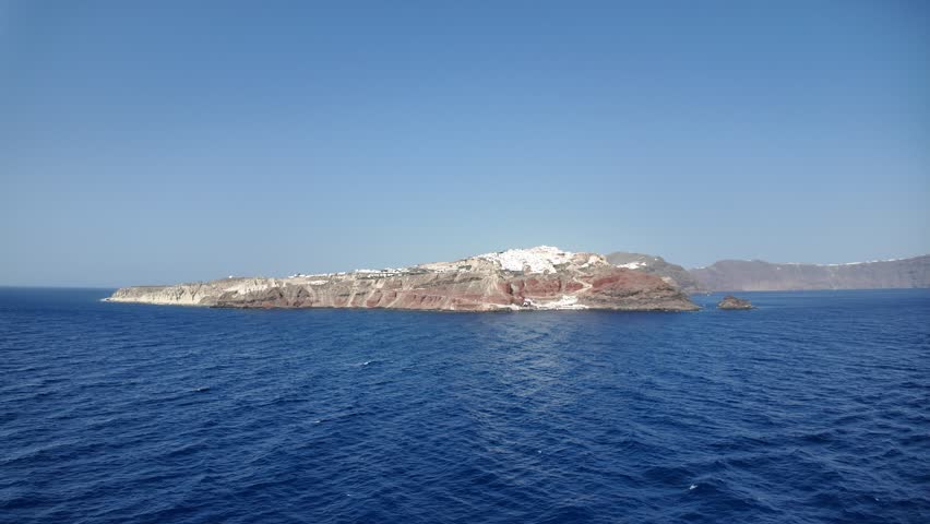 Looking across to Oia on the Greek island of Santorini from a cruise ship leaving the island. There is bright blue water below and a blue sky above.