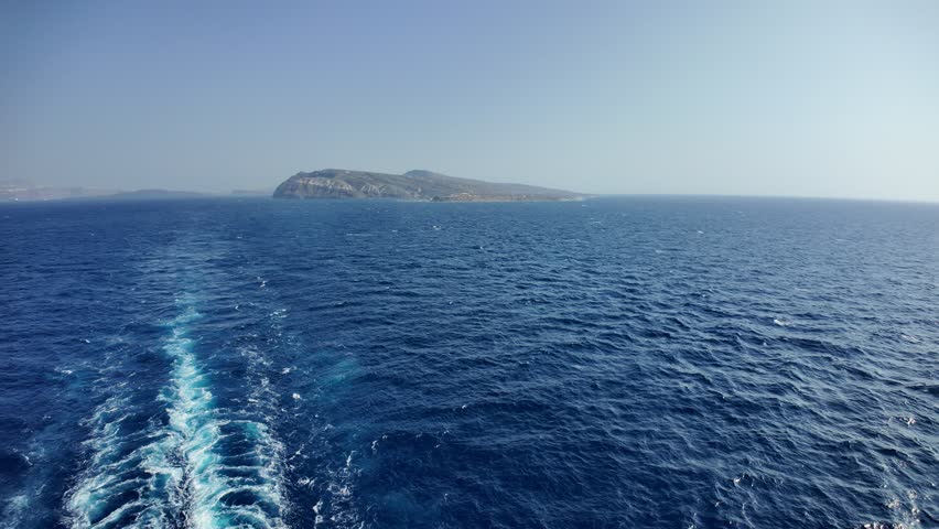 Looking across to the islands of Santorini and Therasia from a cruising ship. There is bright blue water below and a blue sky above.