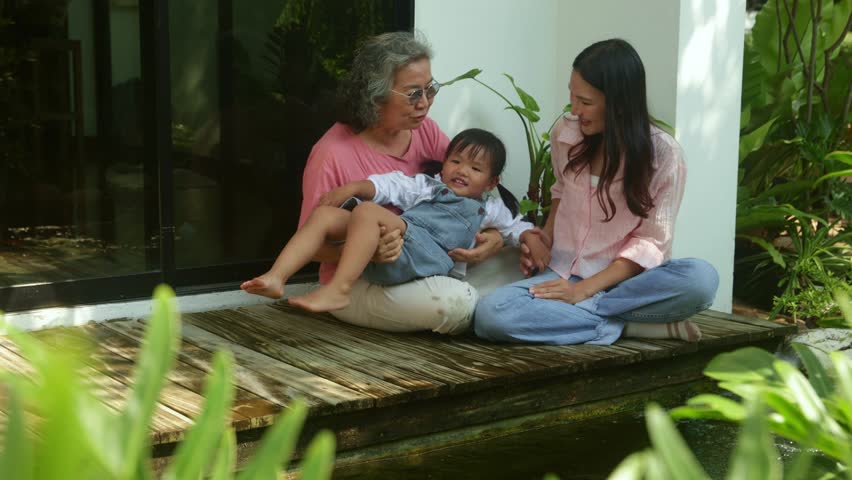 Happy elderly mother her adult daughter and granddaughter sit relaxing and enjoying the peaceful atmosphere beside koi pond enjoying the lush nature of her home : Happy retired women reunited.