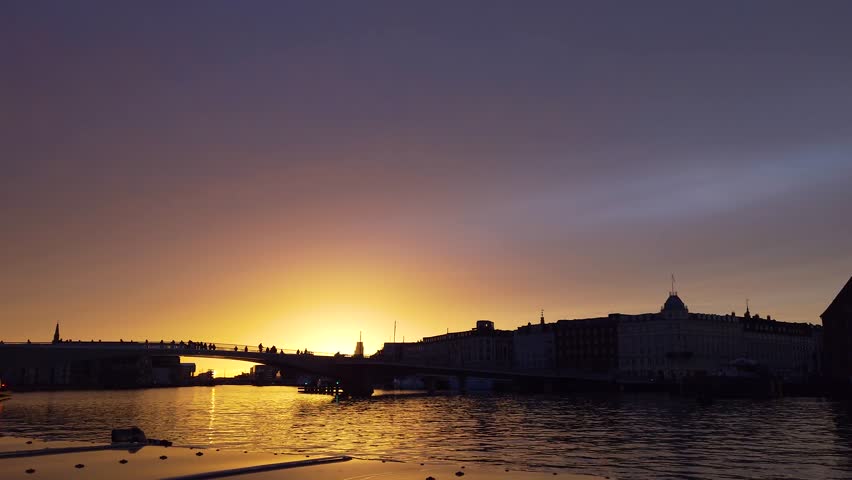Golden sunset silhouettes of bikers crossing a bridge over water in Copenhagen, captured from a moving boat - Denmark