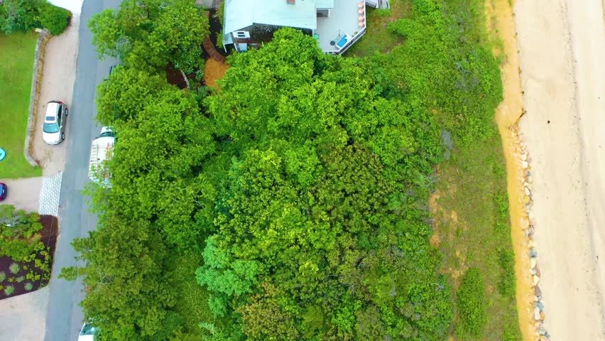 Aerial drone view of Cape Cod beach houses along the shoreline at high tide, framed by marshland and trees, with cloudy skies above and a public parking lot with cars visible near the water’s edge.