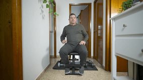Overweight latin senior man over 50 sitting on bench bending sideways to pick up dumbbell before starting workout at home. Realistic indoor fitness and lifestyle scene. - Powered by Shutterstock - Get 15% off with code: PIKWIZARD15