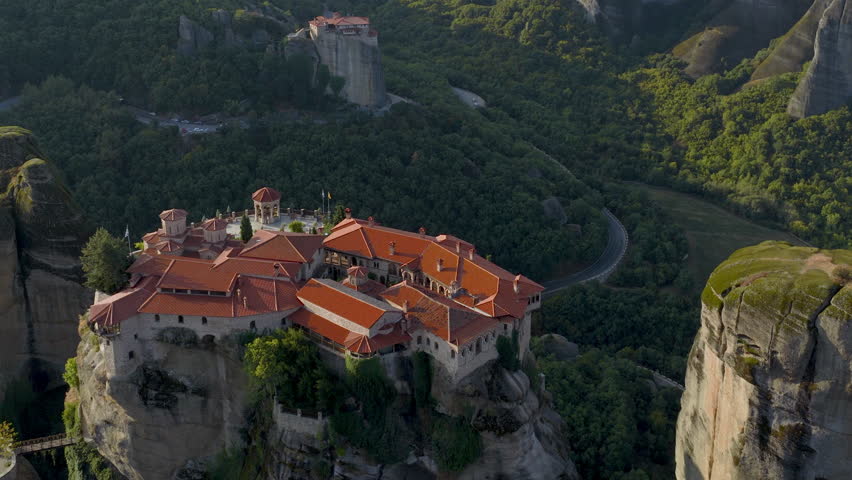 Cinematic aerial view of Meteora monastery in Greece perched on towering cliffs, dramatic rock formations and lush green valley create a breathtaking historic scene