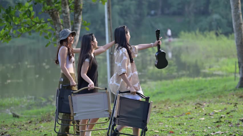 Three women are sitting on lawn chairs and one of them is holding a guitar. The scene is set in a park with a lake in the background. Scene is relaxed and leisurely