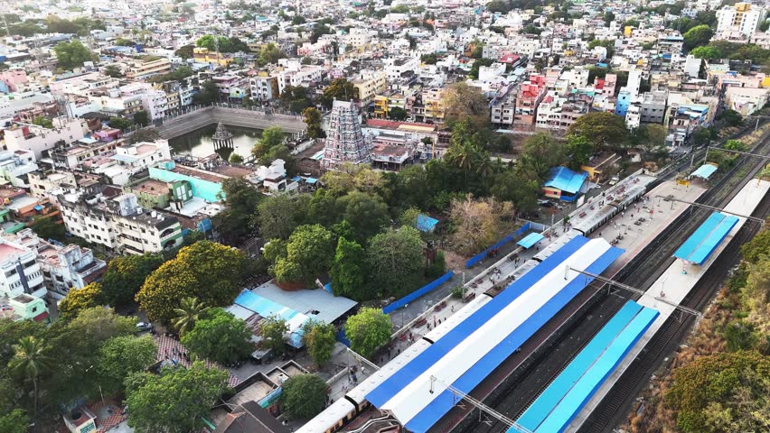 Chennai Saidapet railway station and karneshwar temple. The station has a long foot overbridge with blue-and-white roofing, connecting multiple platforms. A train is visible on one of the tracks.