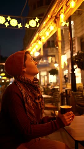 Vertical 4k footage of a young woman in warm clothes sitting at an outdoor cafe at winter night, holding a paper cup of hot coffee with festive city lights in the background