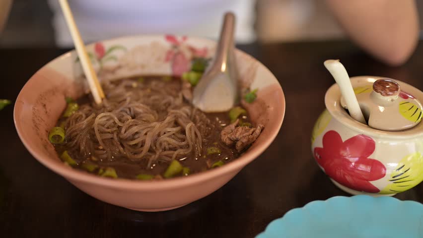 Asian woman eating boat noodles served at restaurant in Thailand adding chili powder to bowl