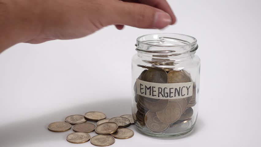 A hand putting gold coins into a jar labeled Emergency. Depositing money into emergency savings jar