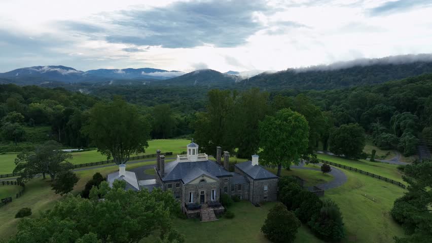 Aerial view of historic Stonehurst Manor in Virginia, surrounded by lush green landscape, trees and Blue Ridge Mountains in background under cloudy sky. Peaceful nature in USA.