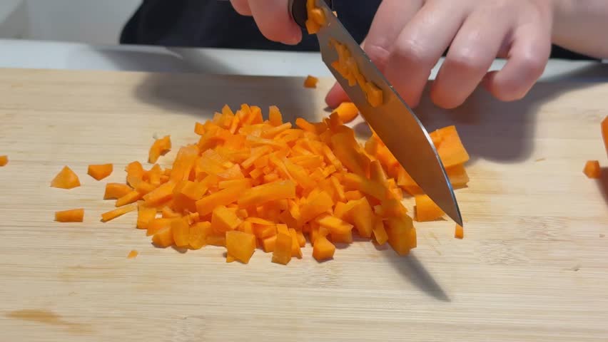 Woman dices fresh orange carrots on a wooden cutting board in the bright daylight of her kitchen