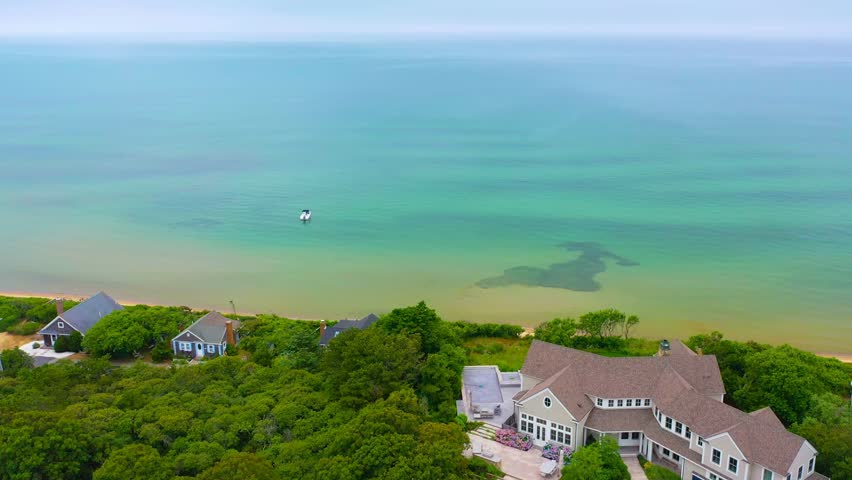 High aerial view of Cape Cod shoreline at high tide, showing beach houses along the coast with a boat visible in the water, framed by cloudy skies and the dramatic atmosphere of the Atlantic.