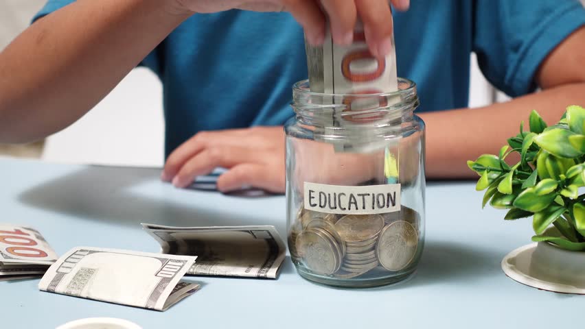 A young girl puts money into a glass jar labeled 'Education,' highlighting the importance of saving for schooling, future learning, and smart financial planning at home