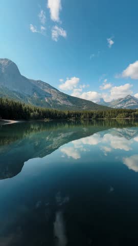 A vertical view of a serene alpine lake in the Canadian Rockies, where crystal-clear reflections mirror snow-capped peaks and drifting clouds