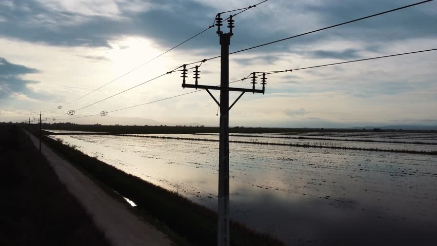 Flooded rice paddies reflect the cloudy sunset sky near power lines and a dirt road
