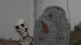 Close-up of a Halloween skeleton with a skull and bones leaning beside a cracked tombstone on a grassy lawn. Spooky outdoor cemetery decoration with tattered fabric, weathered stone, and autumn yard - Powered by Shutterstock - Get 15% off with code: PIKWIZARD15