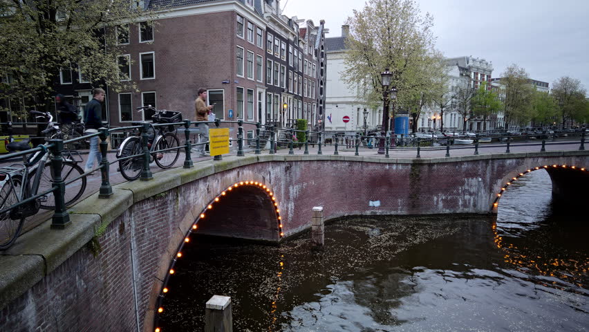 A footage at the intersection of Leidsegracht and Keizersgracht canals in Amsterdam, with antique houses standing at the corner, tour boats cruising under the bridges and street lamps lit up at dusk